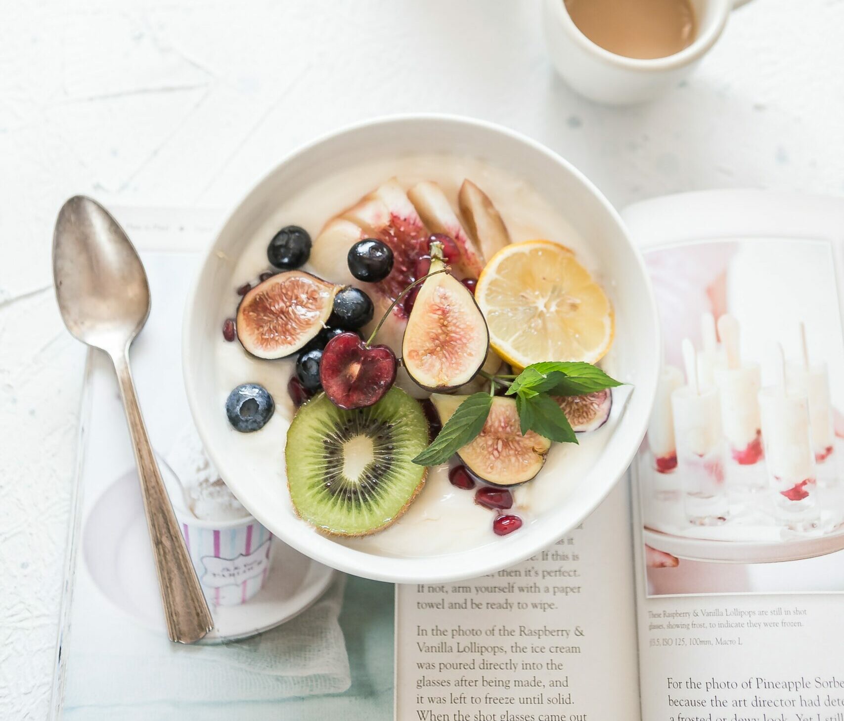 Bowl on white table with yogurt and fresh fruit, coffee in coffee cup, spoon